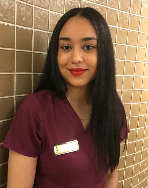 A woman wearing a red lipstick is standing against a tiled wall, posing for the photo with a slight smile.