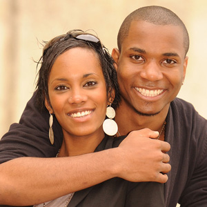 The image shows two individuals embracing each other with smiles, set against a neutral background.